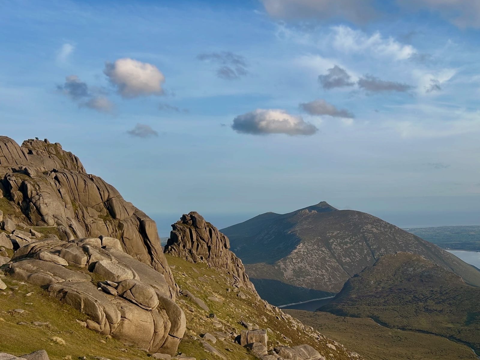 Weathered granite tors on a grassy Mourne slope, with a pointed summit and a reservoir beyond under a blue sky