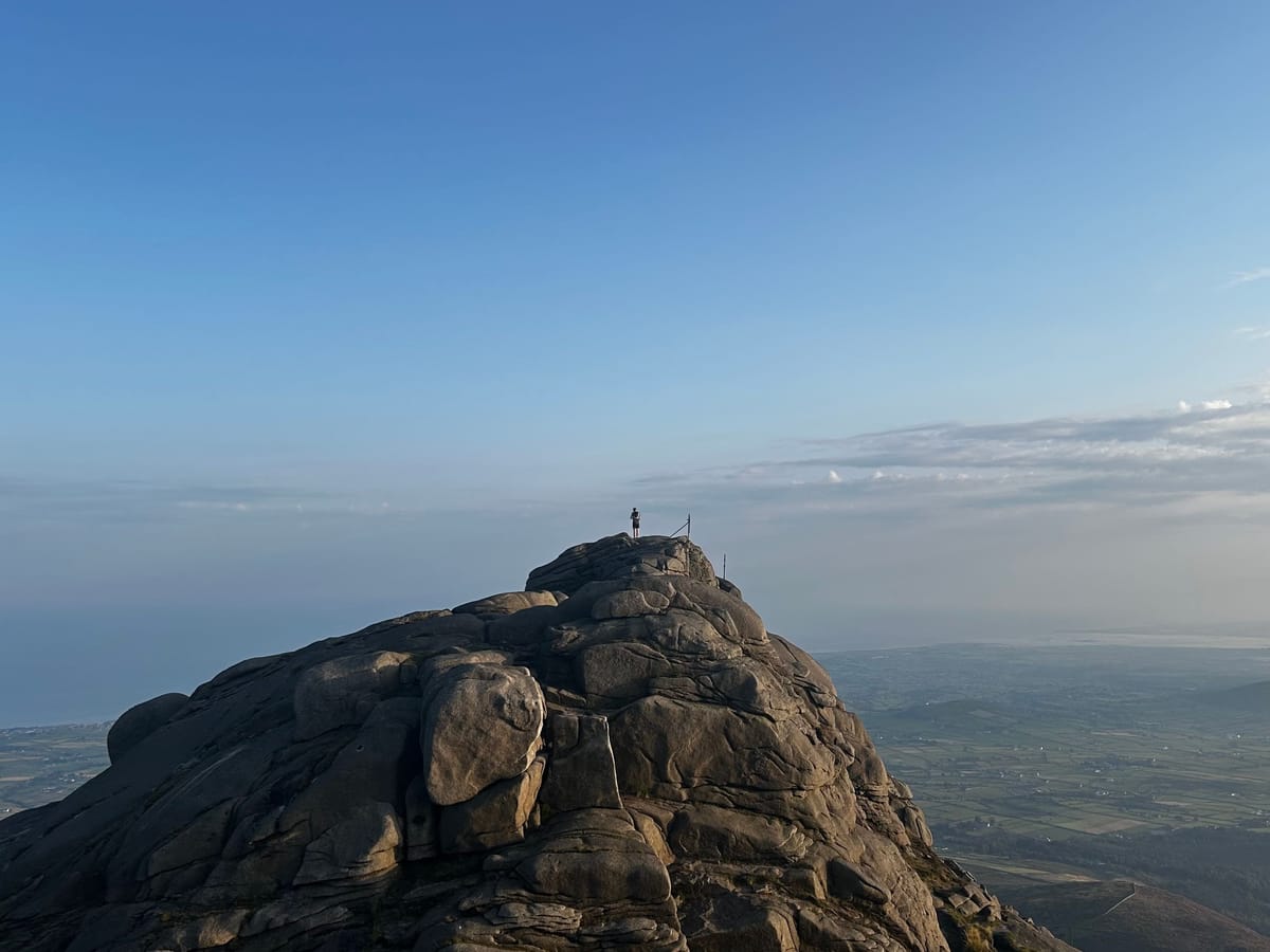 A lone figure stands on top of a huge granite tor in the Mournes, high above farmland and the coast