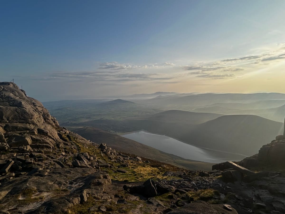 Granite tors frame a reservoir in the valley below, with layered Mourne hills fading into evening haze
